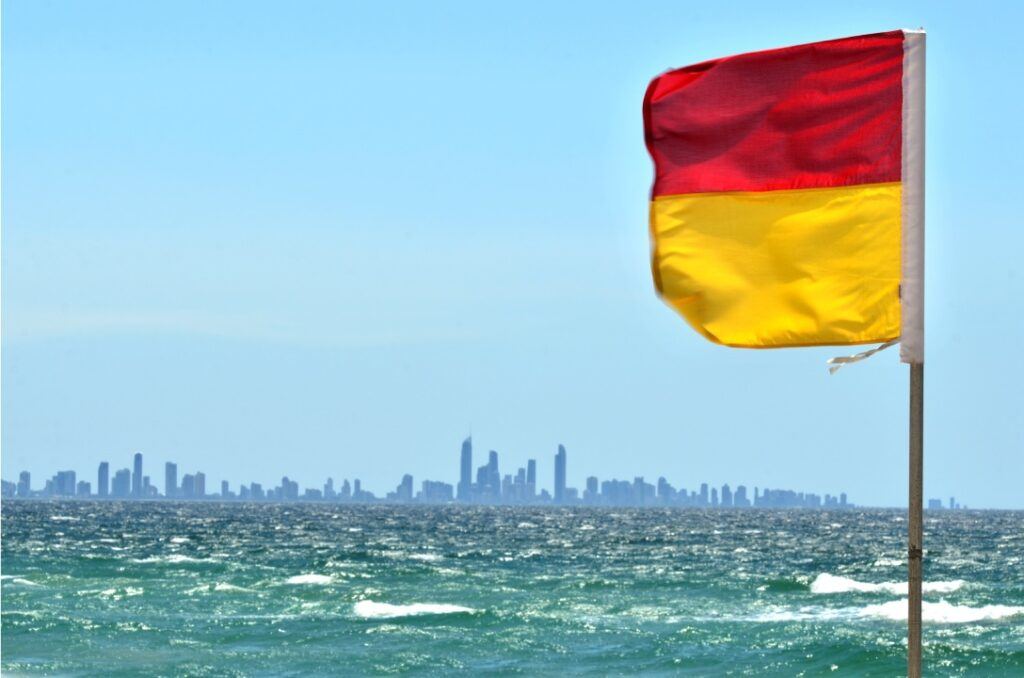 Red and yellow beach safety flag flying near the shoreline, with ocean waves in the foreground and a distant Australian city skyline under a clear blue sky.
