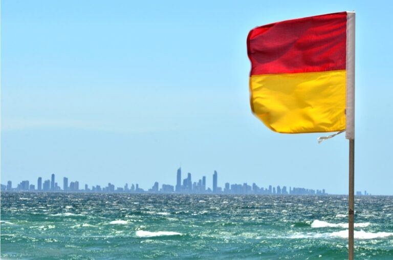 Red and yellow beach safety flag flying near the shoreline, with ocean waves in the foreground and a distant Australian city skyline under a clear blue sky.
