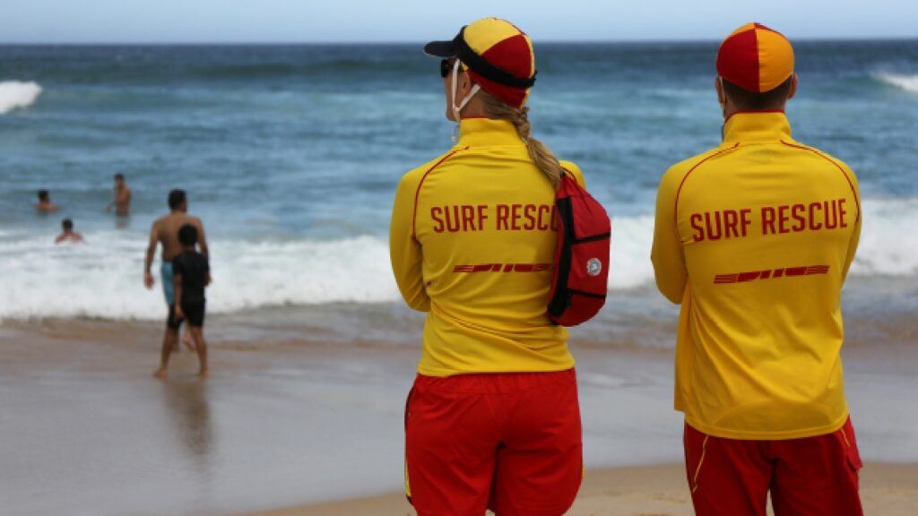 Two Central Coast lifeguards in red and yellow surf rescue uniforms watch over swimmers at the beach, highlighting their vigilance and role in public safety.