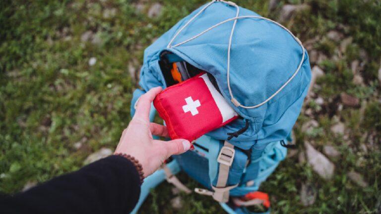 A person pulling out a red survival handy first aid kit from a blue hiking backpack on a grassy trail, prepared for emergencies while outdoors.