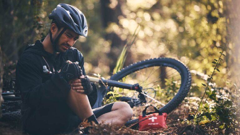 Injured mountain biker sitting on a forest trail, treating his knee with a visible Mountain Biking First Aid Kit beside his fallen bike.