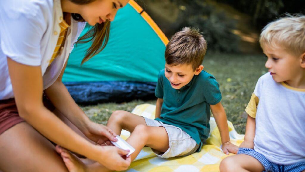 Mother using a family first aid kit to treat her child’s scraped knee while camping outdoors with another child watching.